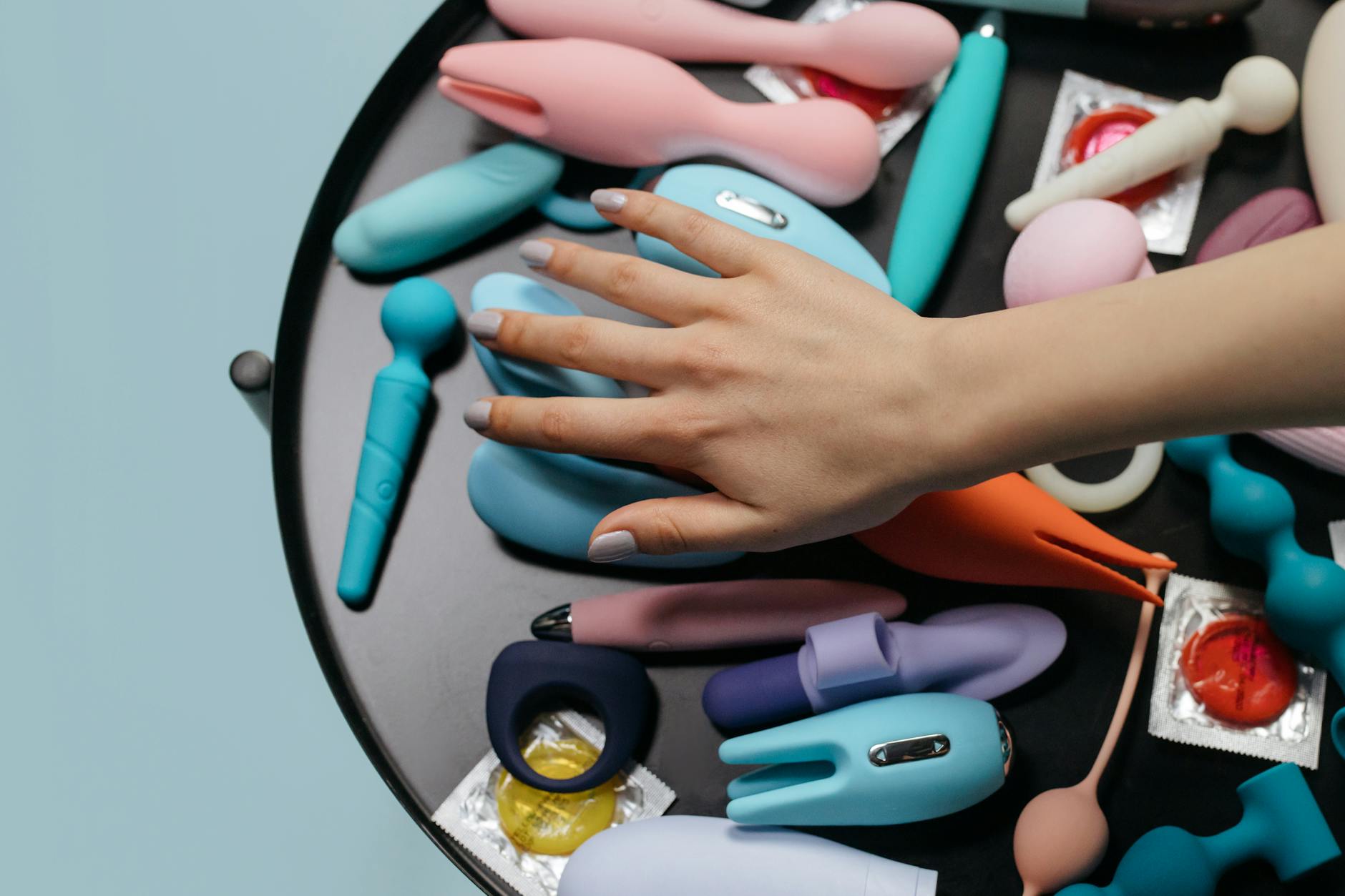 A hand reaching over a variety of colorful clitoral vibrators and adult toys arranged on a table