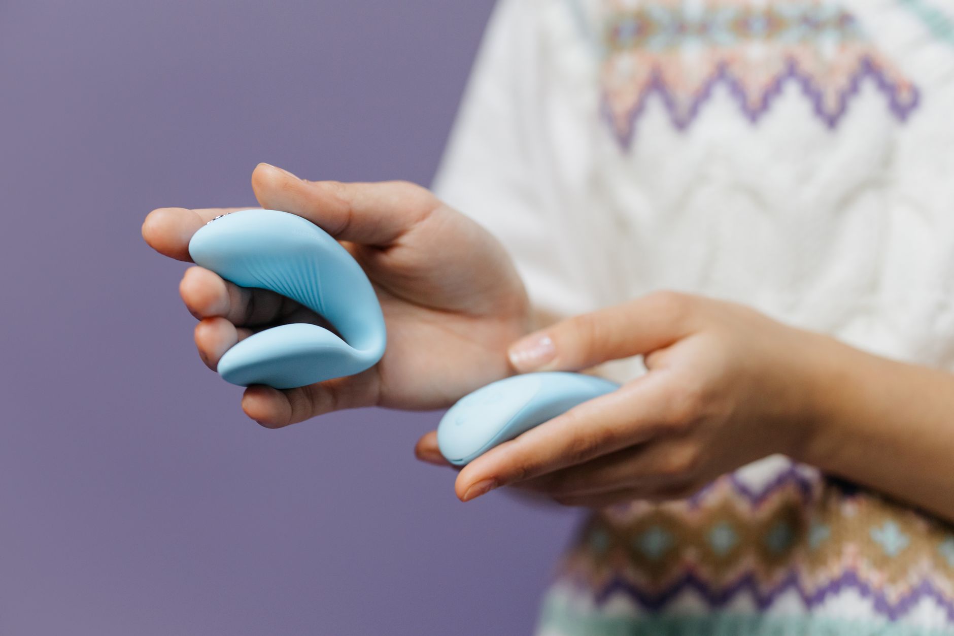 Mãos segurando um vibrador silicone azul contra fundo roxo.
