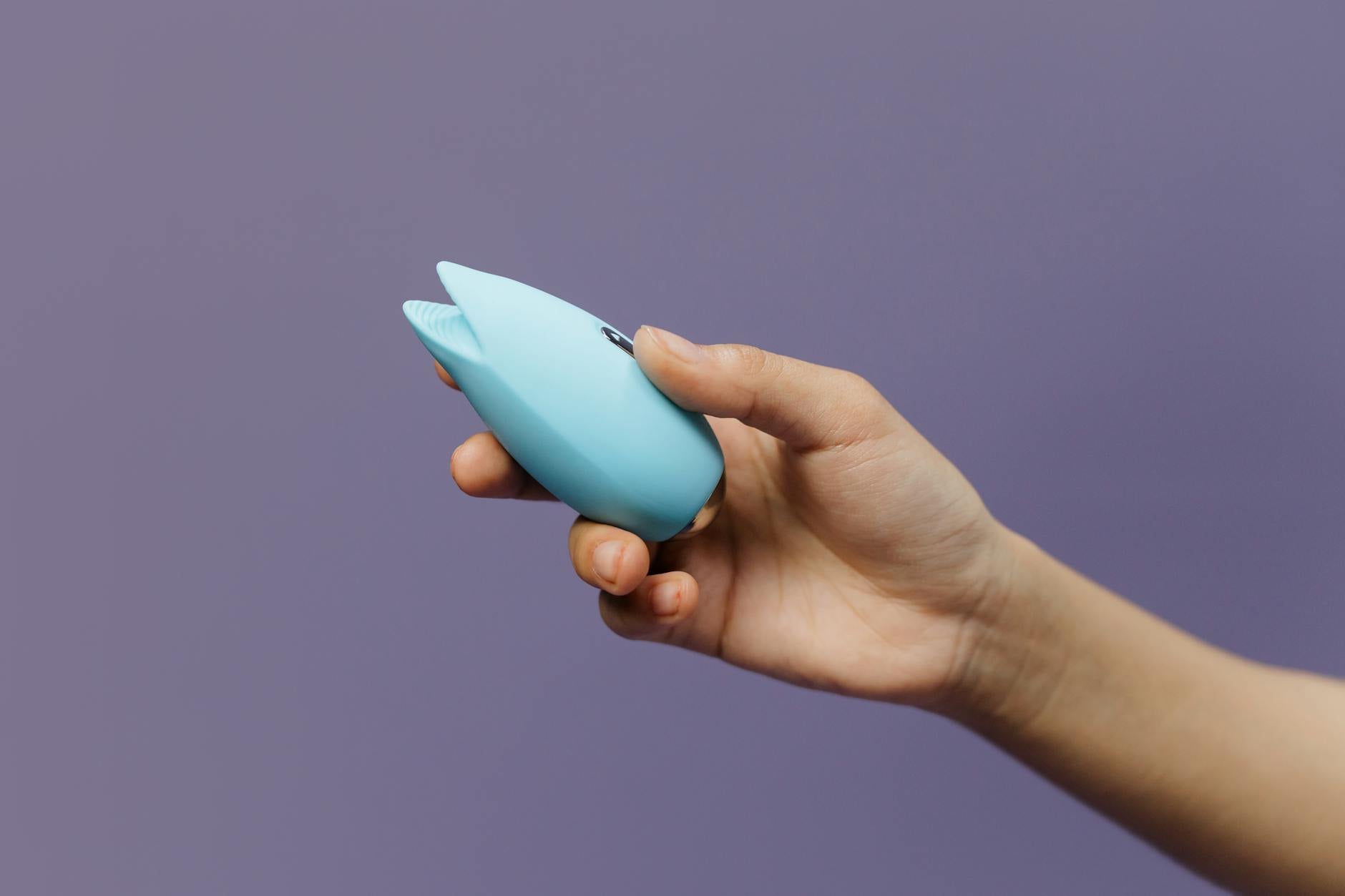 A blue clitoral vibrator held gently in hand against a purple background, symbolizing self-care and reconnection with pleasure.